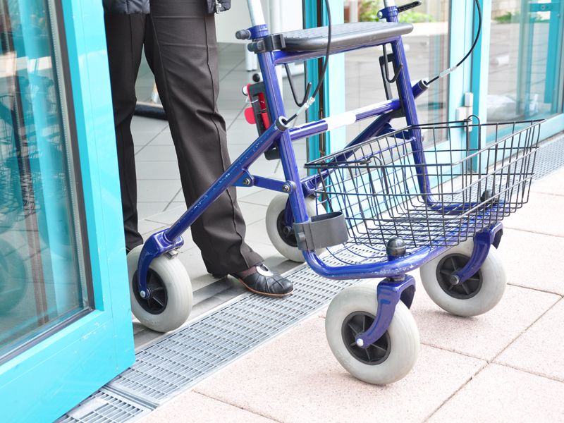 Eine Person mit Rollator überquert eine barrierefreie, schwellenlose Tür zu einem Gebäude mit blauen Rahmen.